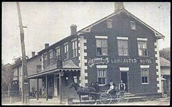 A historic black‑and‑white photograph of the original Lancaster Hotel, showing a horse‑drawn carriage in front of the two‑story brick building in the early 1900s; this same print hangs inside The Lancaster Smokehouse as part of its heritage display.